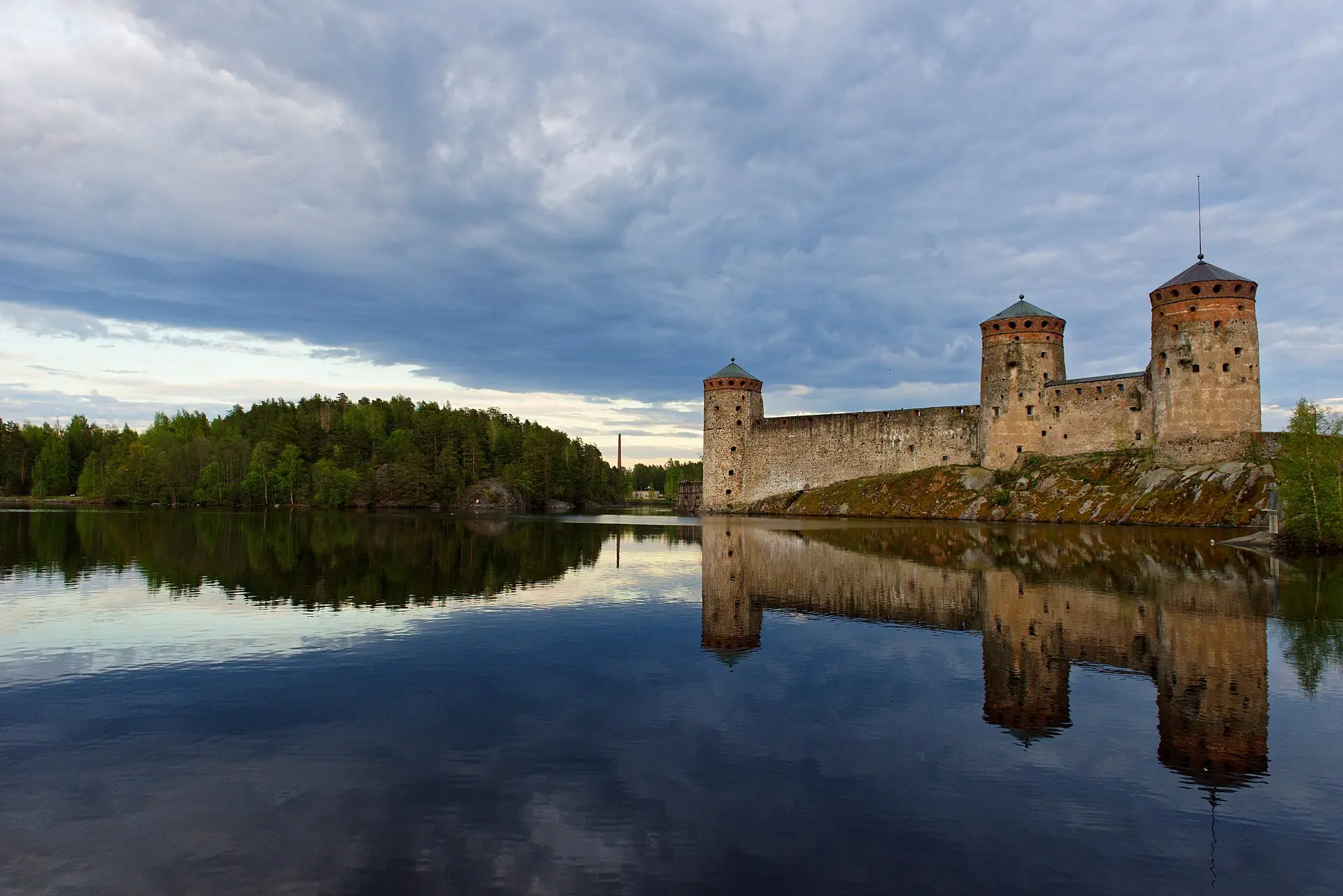 Burg Olavinlinna in Savonlinna: Steinmauer mit drei runden Türmen auf einem Felsen, die sich im ruhigen Wasser spiegelt