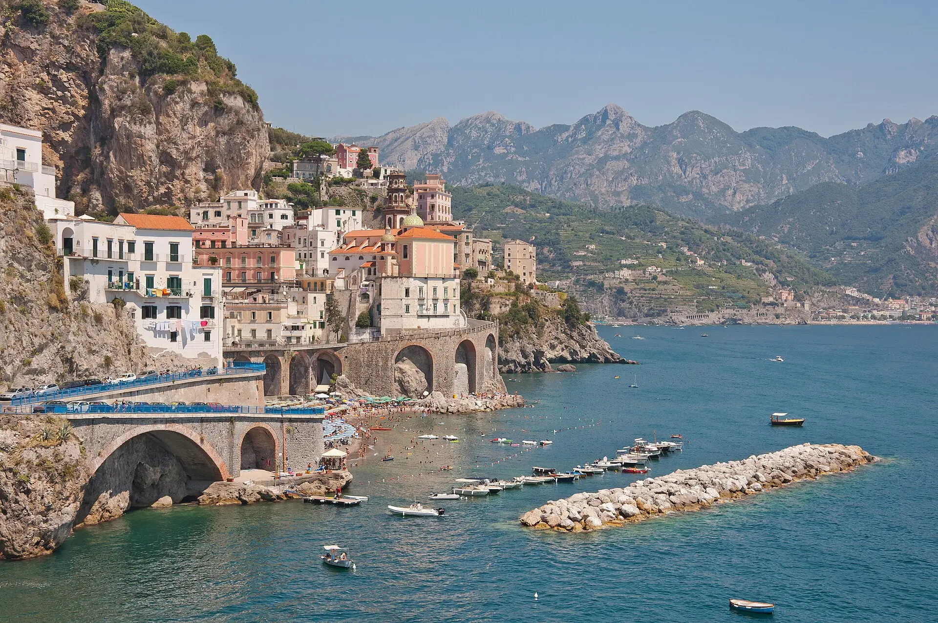 Atrani an der Amalfiküste: helle, gestaffelte Häuser am Fels, eine Straße auf Steinbögen, ein Strand mit Sonnenschirmen und Booten