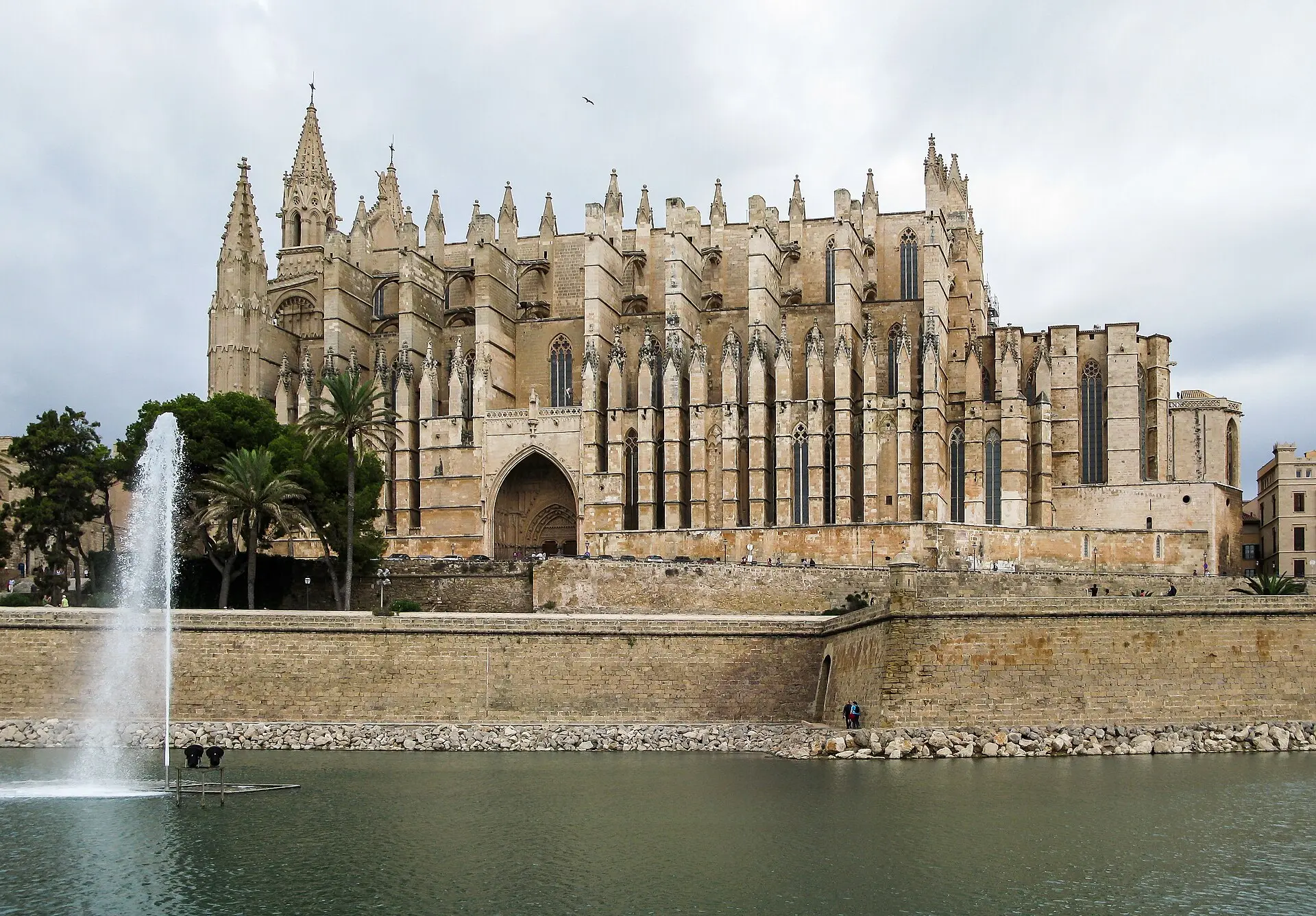 Südseite der Kathedrale La Seu in Palma mit Strebewerk und Portal, davor die Stadtmauer und ein Wasserbecken mit Fontäne