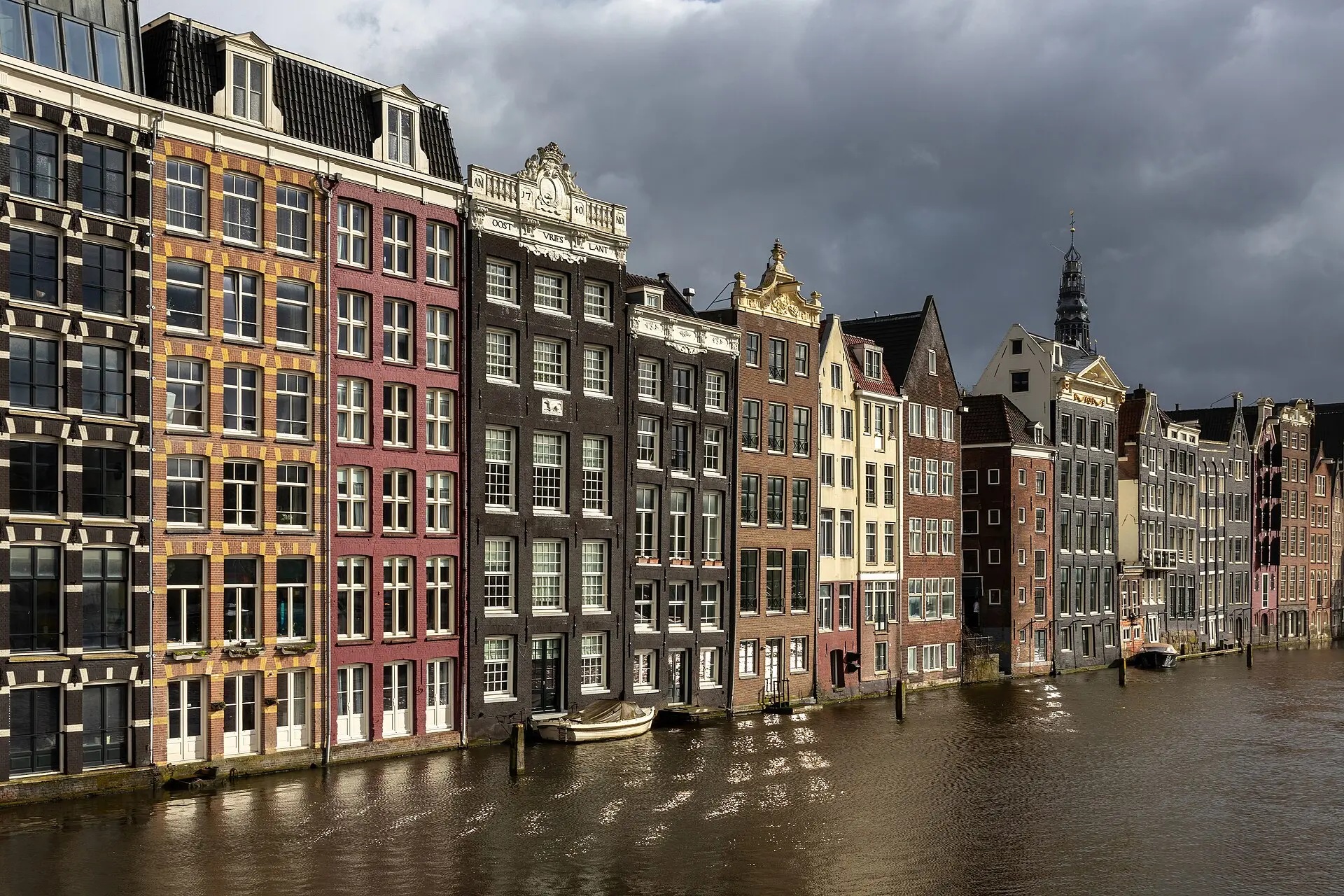 Schmale Giebelhäuser in Schwarz, Rot, Gelb und Grau am Wasser des Damrak, dahinter der Turm der Oude Kerk unter dunklen Wolken