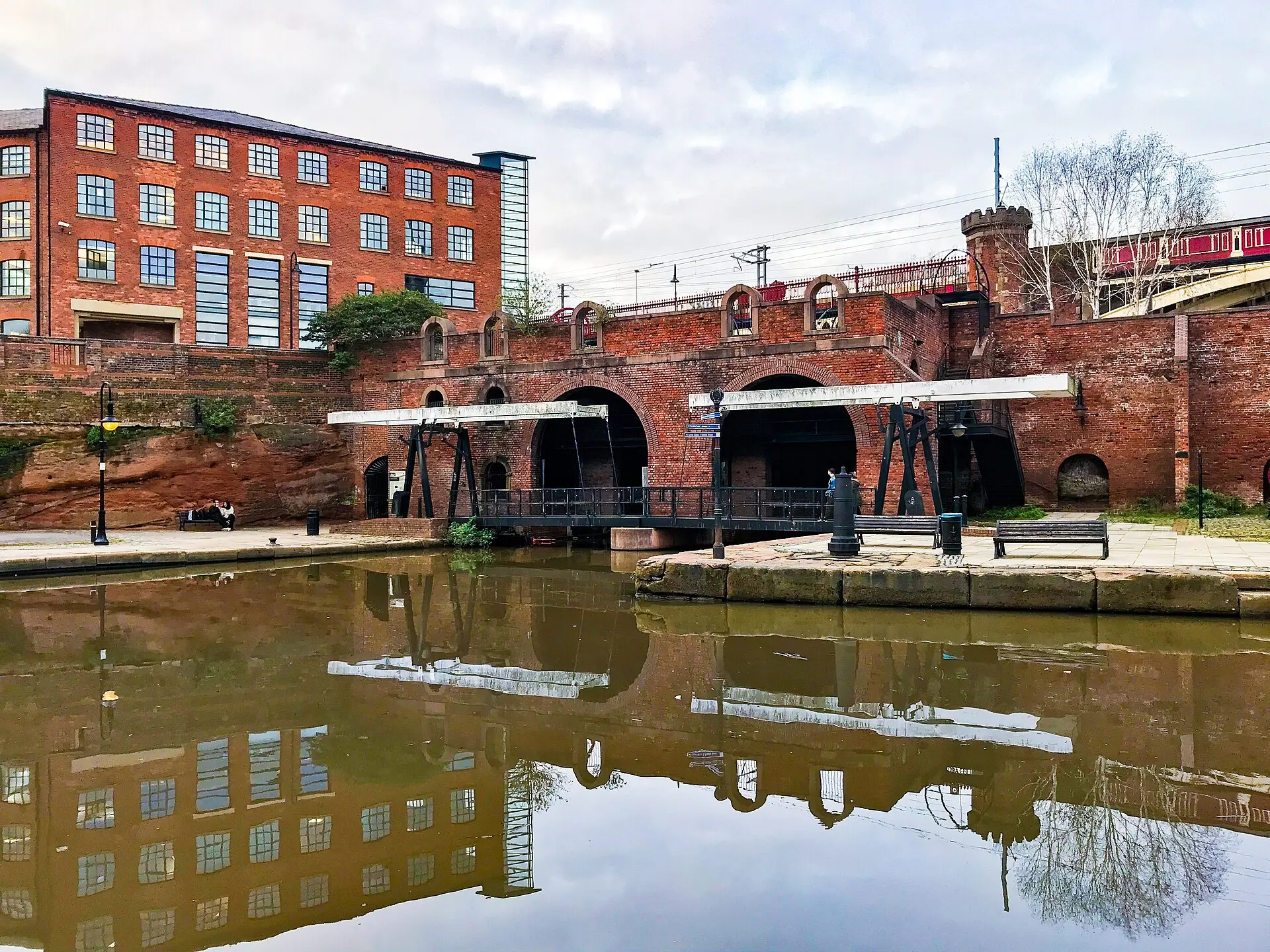 Backstein-Lagerhaus, schwarze Brücke und Eisenbahnviadukt mit rotem Zug spiegeln sich im Kanalbecken von Castlefield