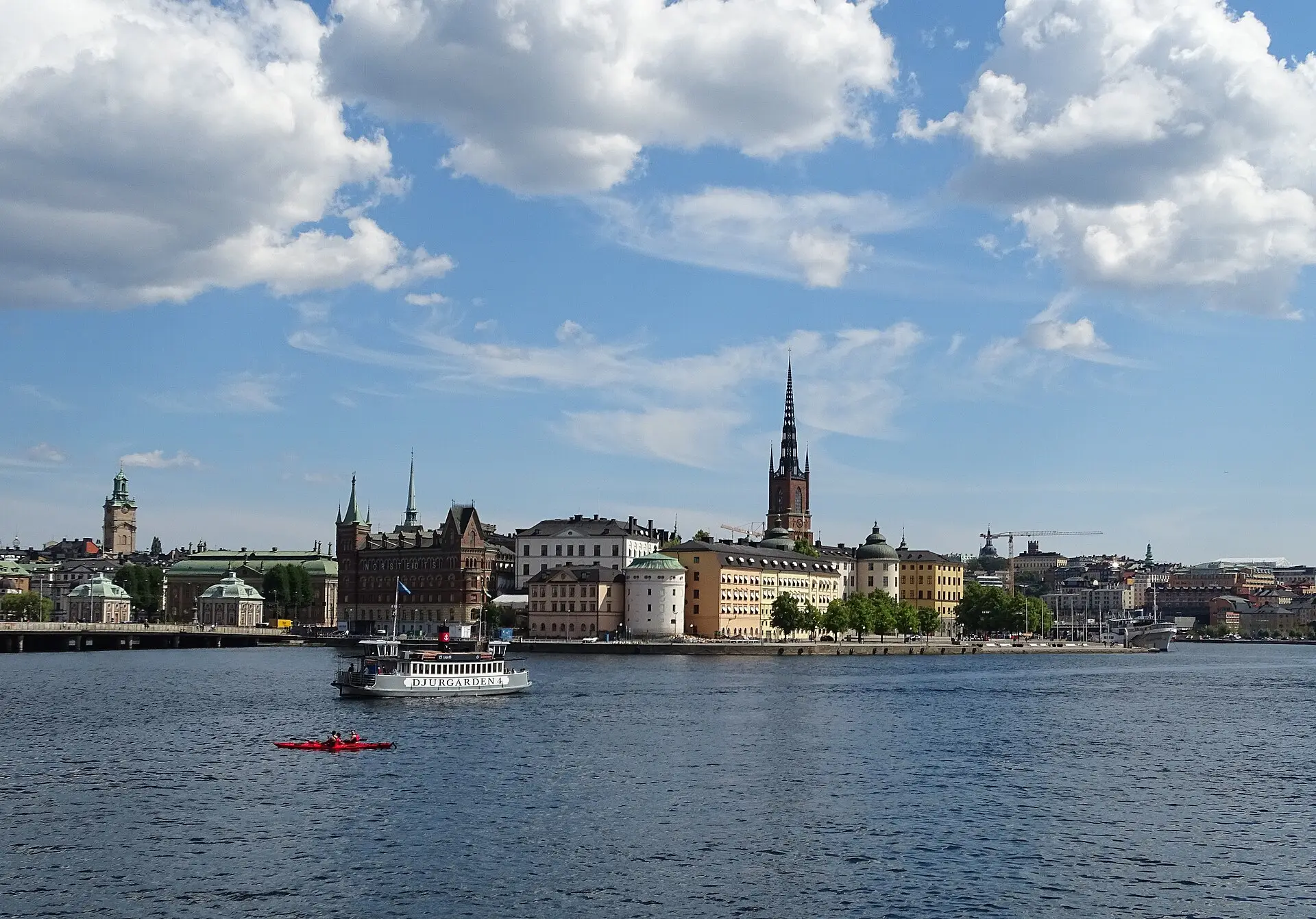 Blick übers Wasser auf Riddarholmen mit dem dunklen Spitzturm der Riddarholmskyrkan, davor eine weiße Fähre und ein rotes Kajak