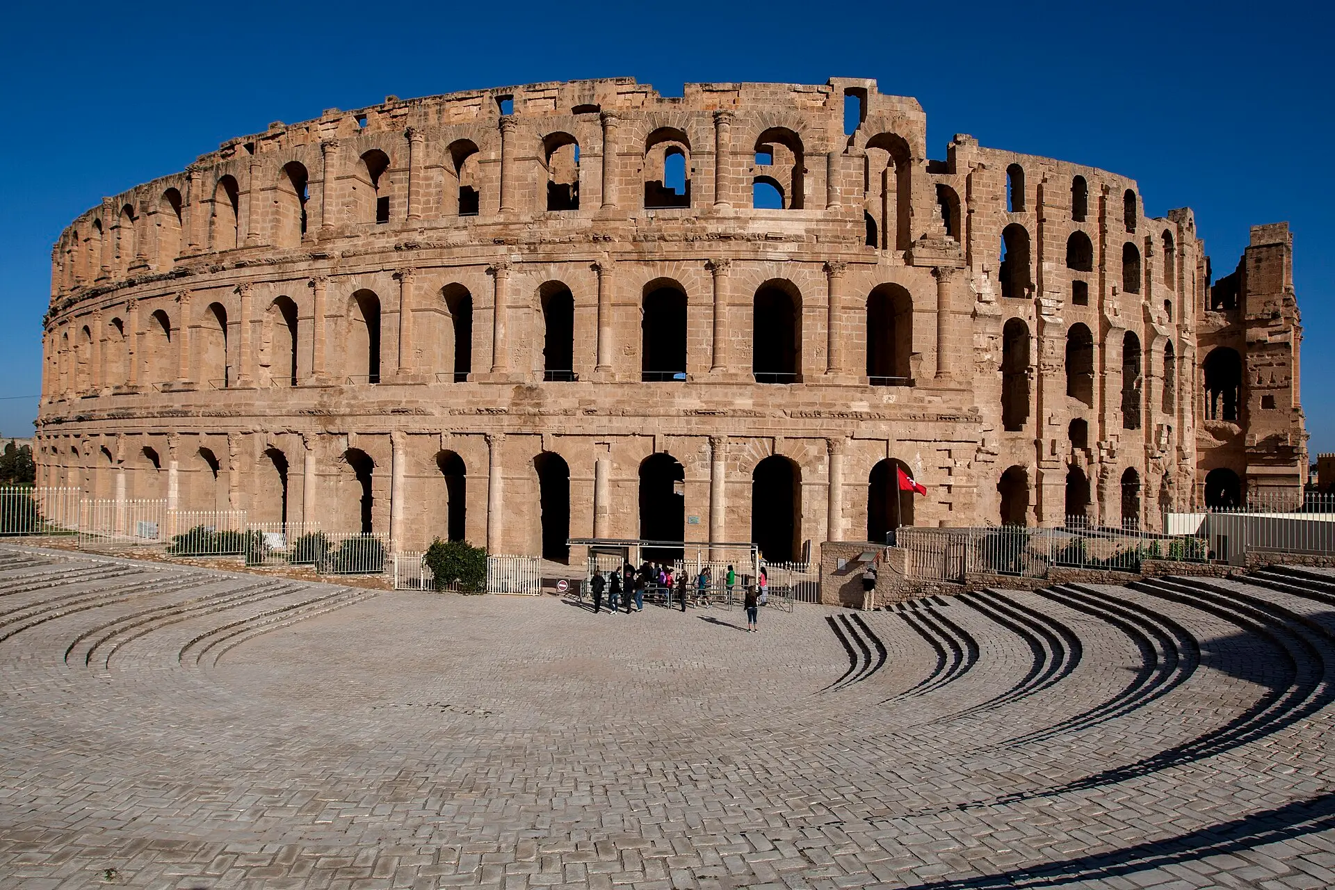Amphitheater von El Jem mit drei Bogenreihen und eingestürztem Abschnitt rechts, davor ein gepflasterter Platz mit kleiner Besuchergruppe