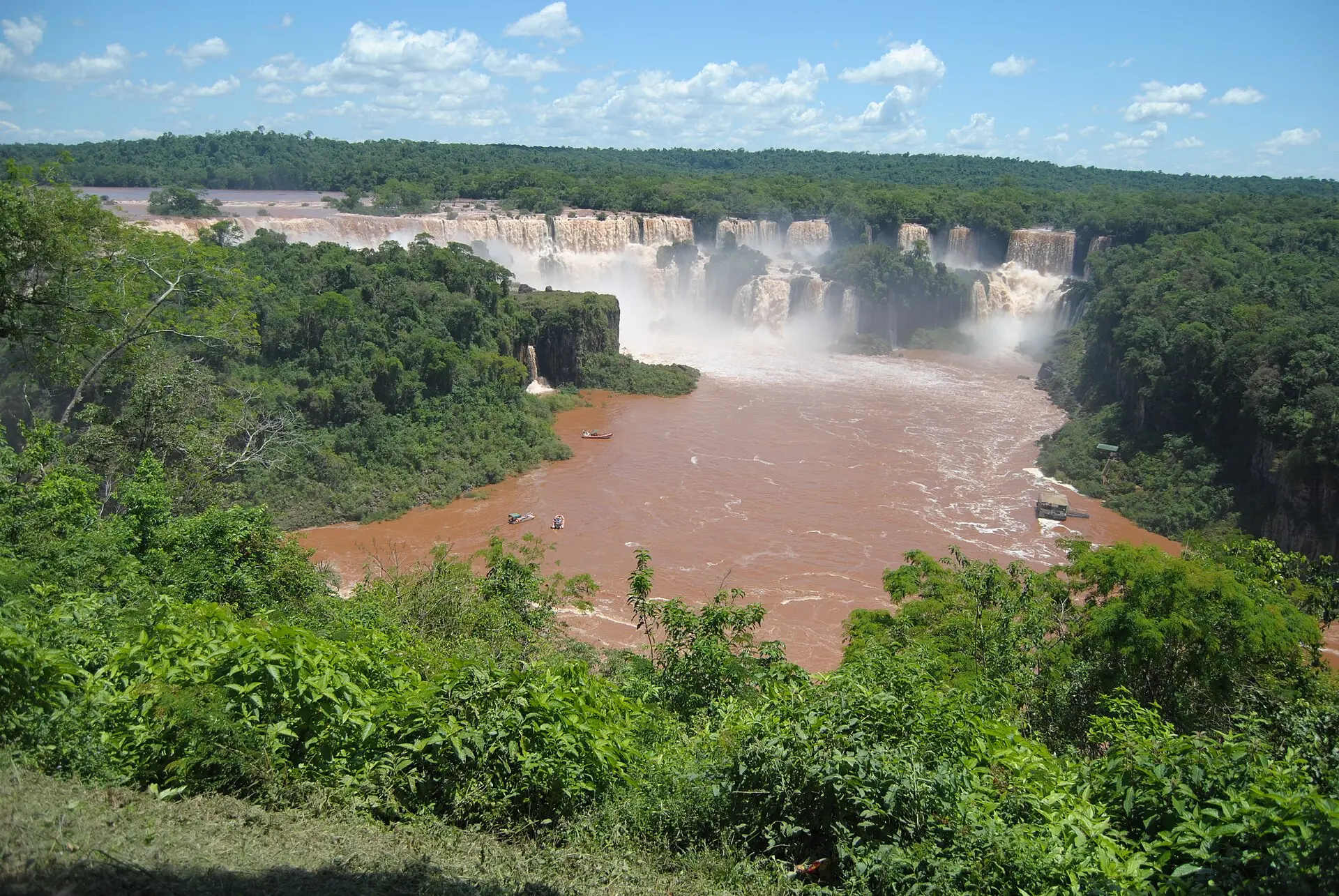 Breite Kette der Iguaçu-Wasserfälle mit Gischt, davor ein brauner Fluss mit kleinen Booten, ringsum dichter grüner Wald