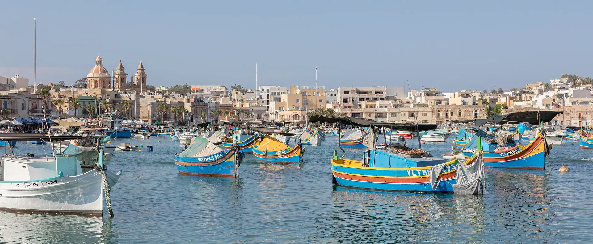 Bunt bemalte Luzzu-Fischerboote im Hafen von Marsaxlokk, dahinter die Uferzeile mit Palmen und einer Kirche mit zwei Türmen