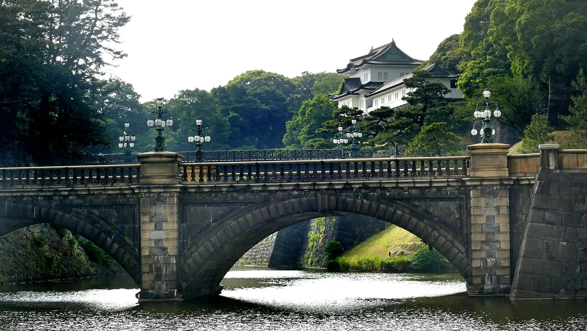 Zweibogige Steinbrücke mit Laternen über den Graben des Kaiserpalasts, dahinter ein weißer Wachturm an einem bewaldeten Hang