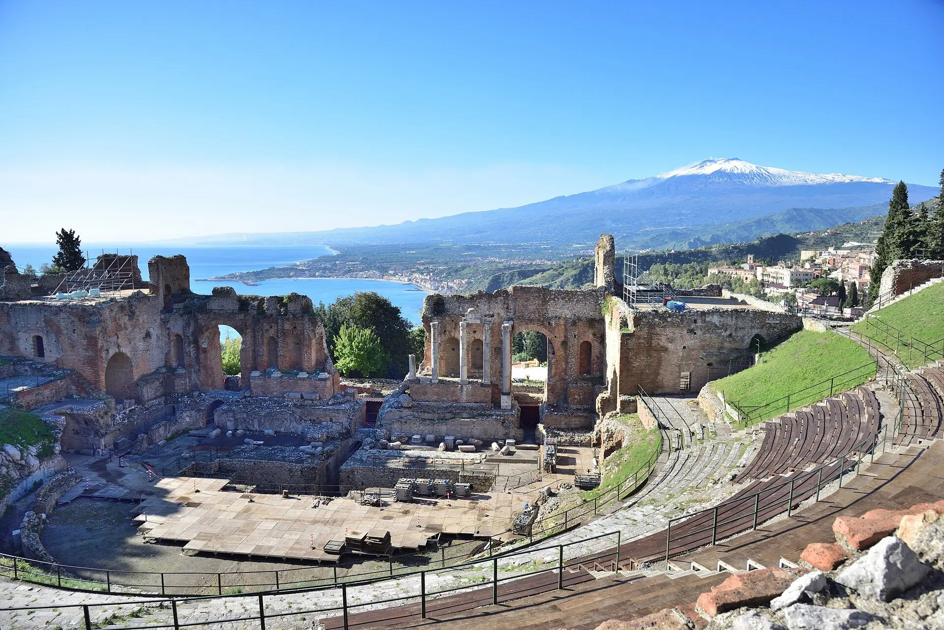 Backsteinruinen des Bühnenhauses im antiken Theater von Taormina, davor ein Bühnenpodest, dahinter Küste, Meer und der verschneite Ätna