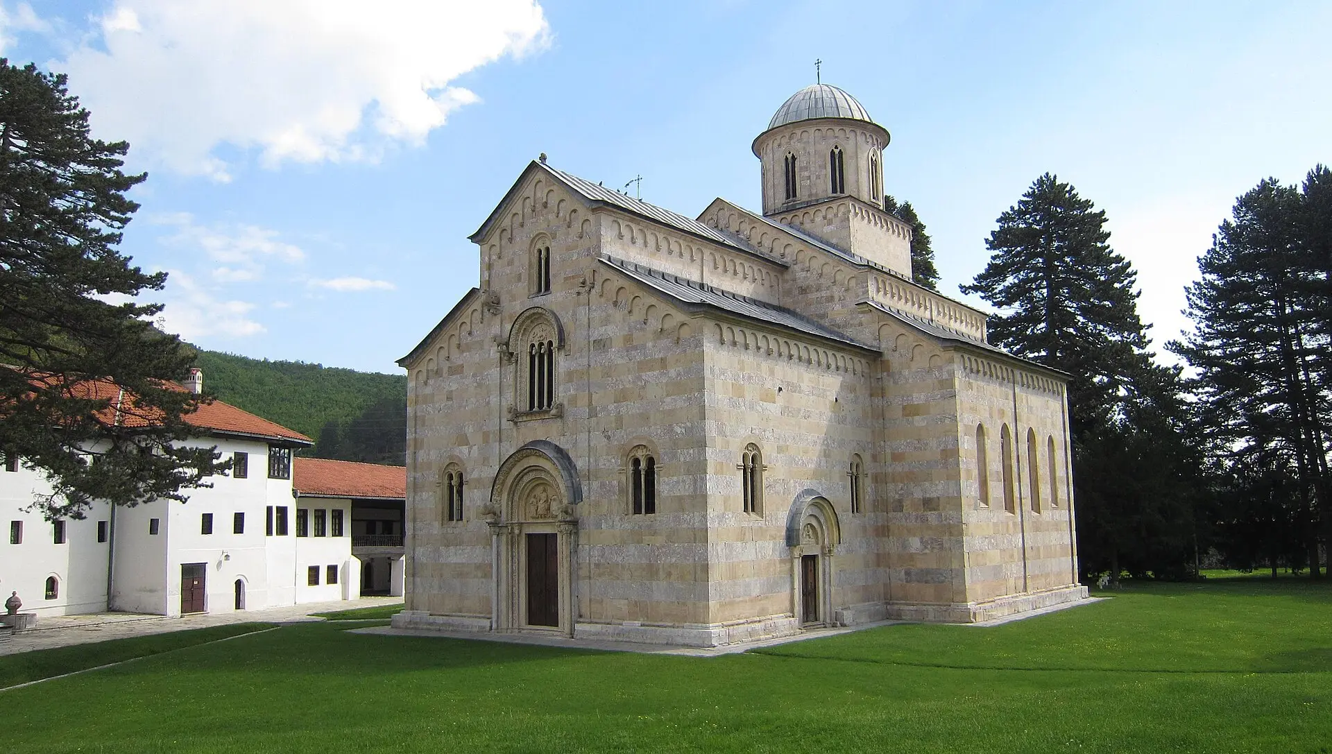 Klosterkirche Visoki Dečani mit Fassade aus hellen und rötlich-grauen Steinbändern, Kuppel und gemähter Wiese