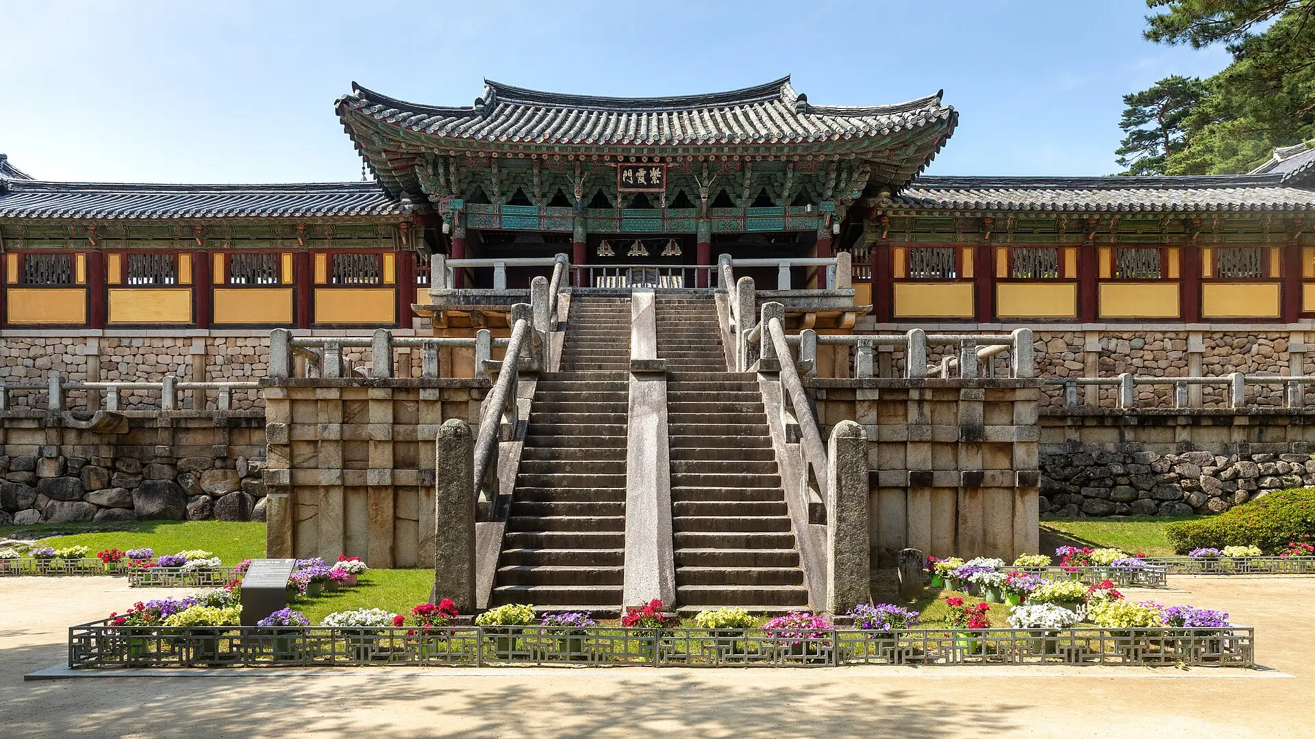 Steinerne Doppeltreppen des Tempels Bulguksa in Gyeongju führen zum Tor Jahamun mit bemaltem Holzdach, davor ein niedriges Blumenbeet