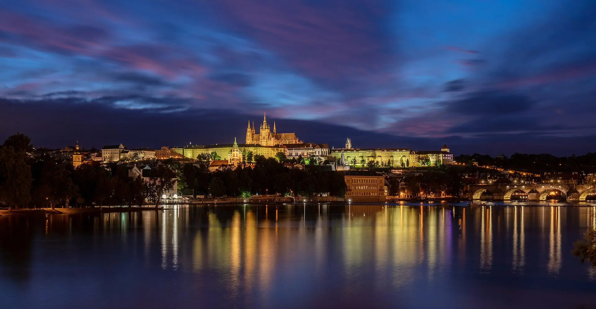 Beleuchtete Prager Burg mit Veitsdom zur blauen Stunde, gespiegelt in der Moldau, rechts die Karlsbrücke
