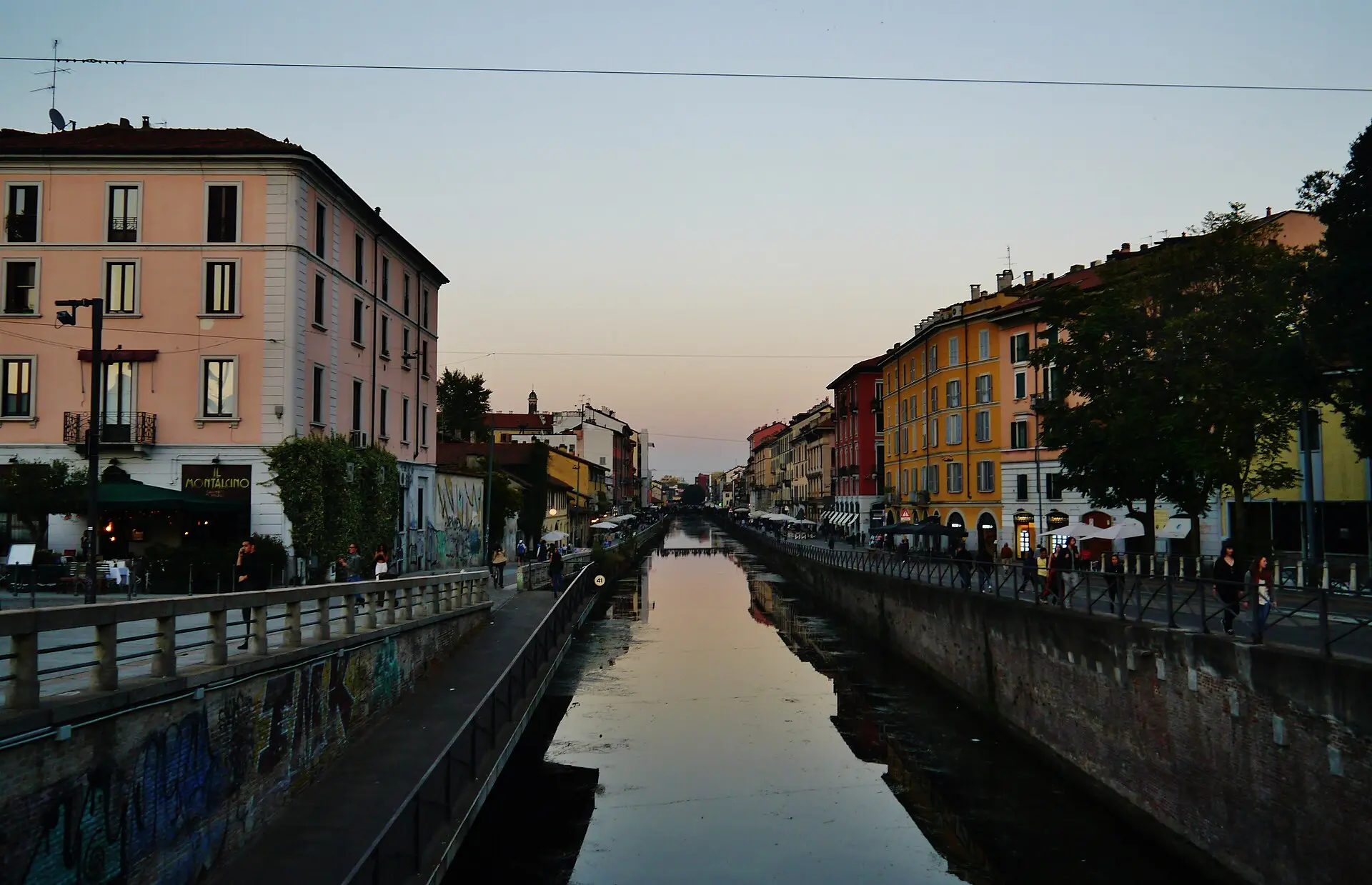 Naviglio Grande am Abend, links ein rosa Eckhaus, rechts gelbe und orange Häuser, Passanten an beiden Ufern, Spiegelungen im Wasser