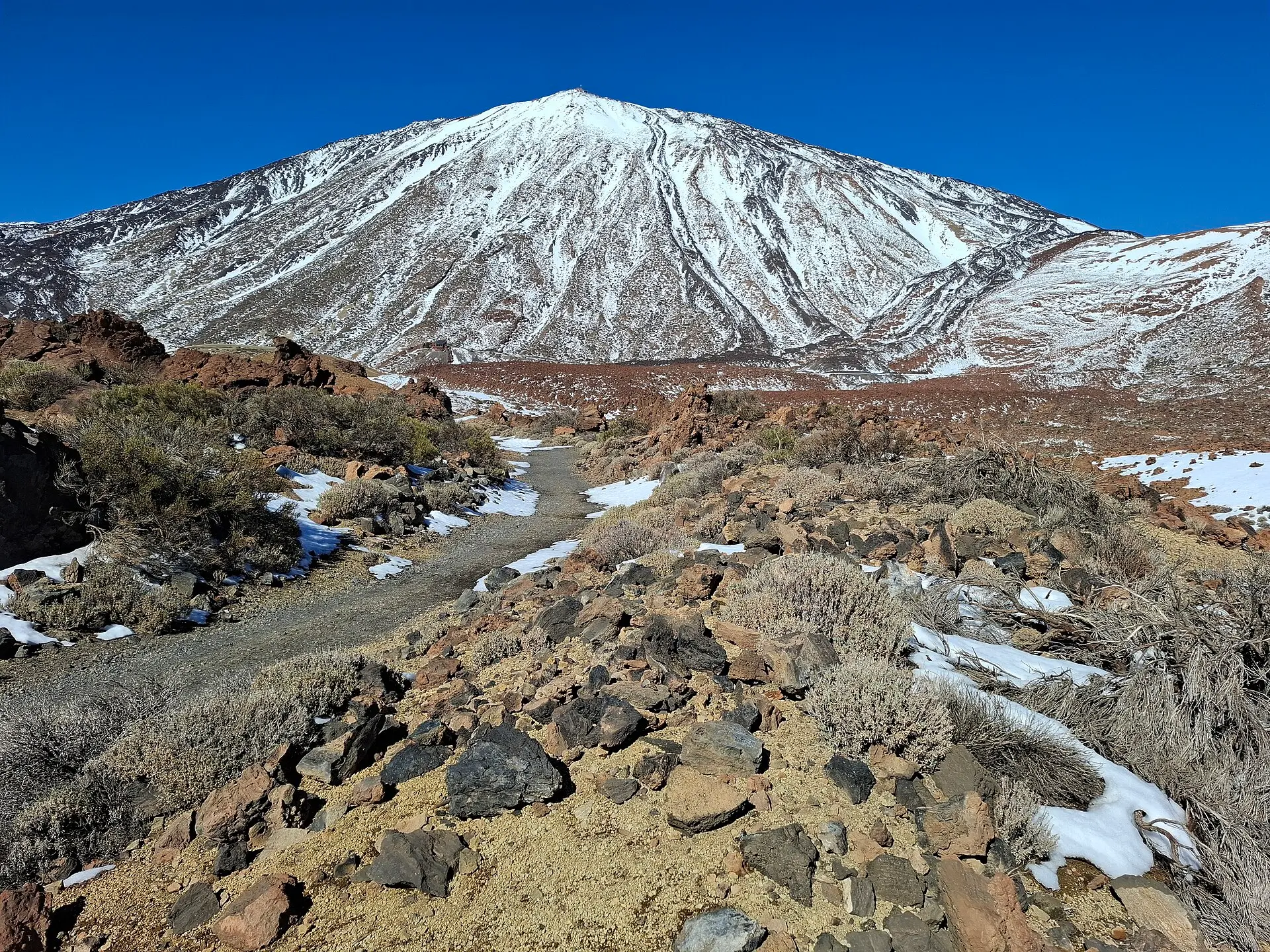 Verschneiter Gipfel des Teide vor blauem Himmel, im Vordergrund ein Wanderweg durch Lavageröll im Nationalpark