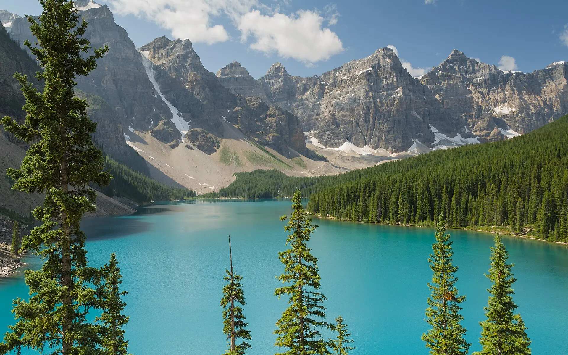 Türkisfarbener Moraine Lake im Banff-Nationalpark, rechts Nadelwald, dahinter graue Felsgipfel mit Schneeresten unter blauem Himmel
