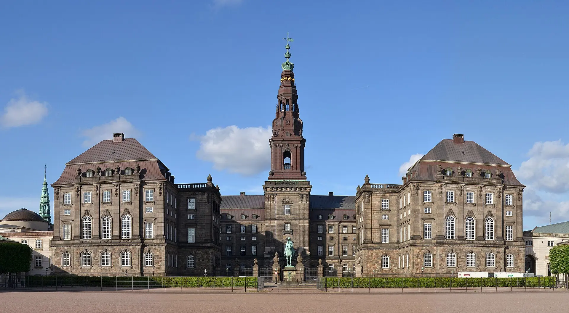 Schloss Christiansborg von vorn mit hohem Mittelturm, davor ein Reiterstandbild und ein leerer Vorplatz unter blauem Himmel