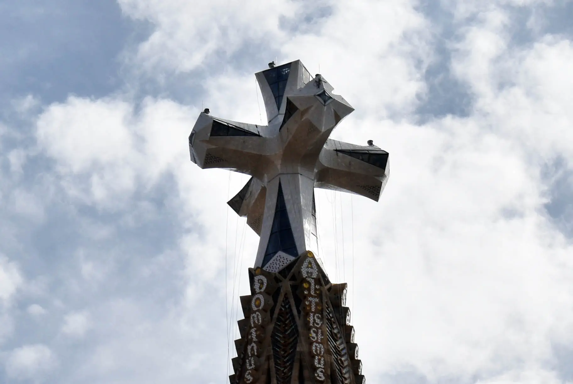 Mit Glas und weißer Keramik verkleidetes Kreuz auf der Spitze des Jesus-Christus-Turms der Sagrada Família vor bewölktem Himmel