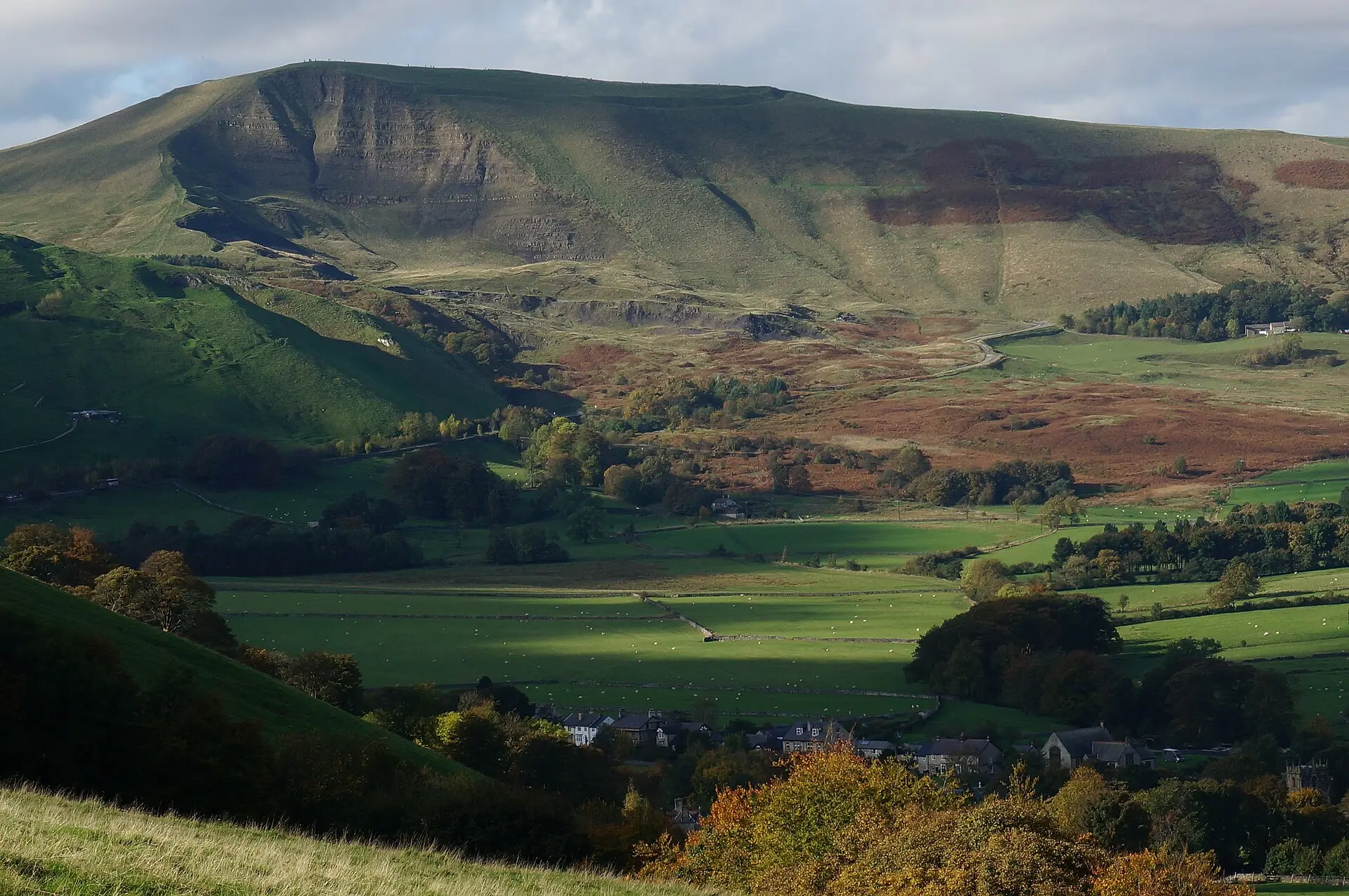 Herbstlich gefärbte Bäume und Dächer von Castleton, dahinter grüne Weiden und der Mam Tor mit steiler, kahler Abbruchkante