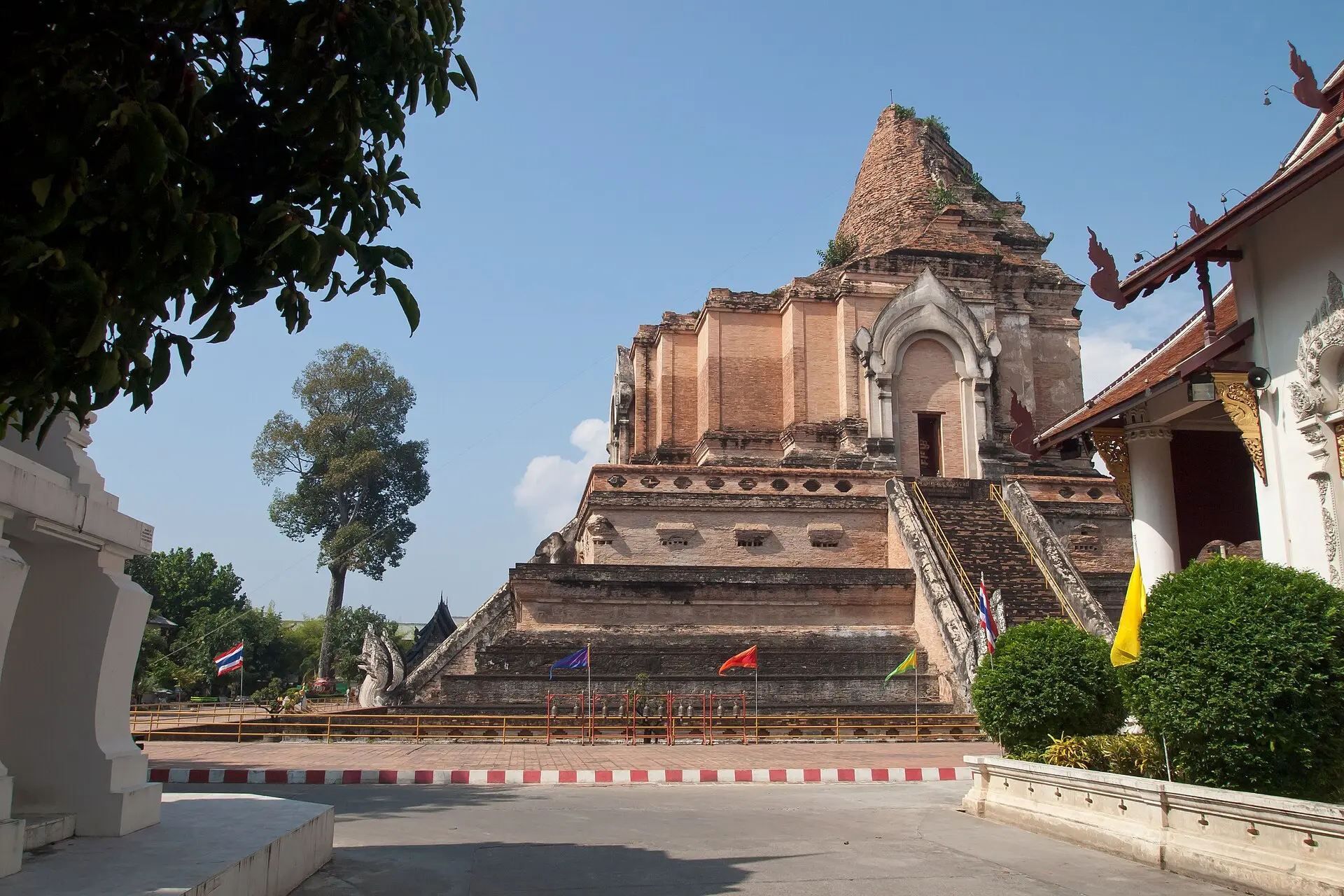 Oben eingestürzter Ziegel-Chedi des Wat Chedi Luang mit Naga-Treppe und kleinen Fahnen, rechts ein Tempelgiebel, blauer Himmel
