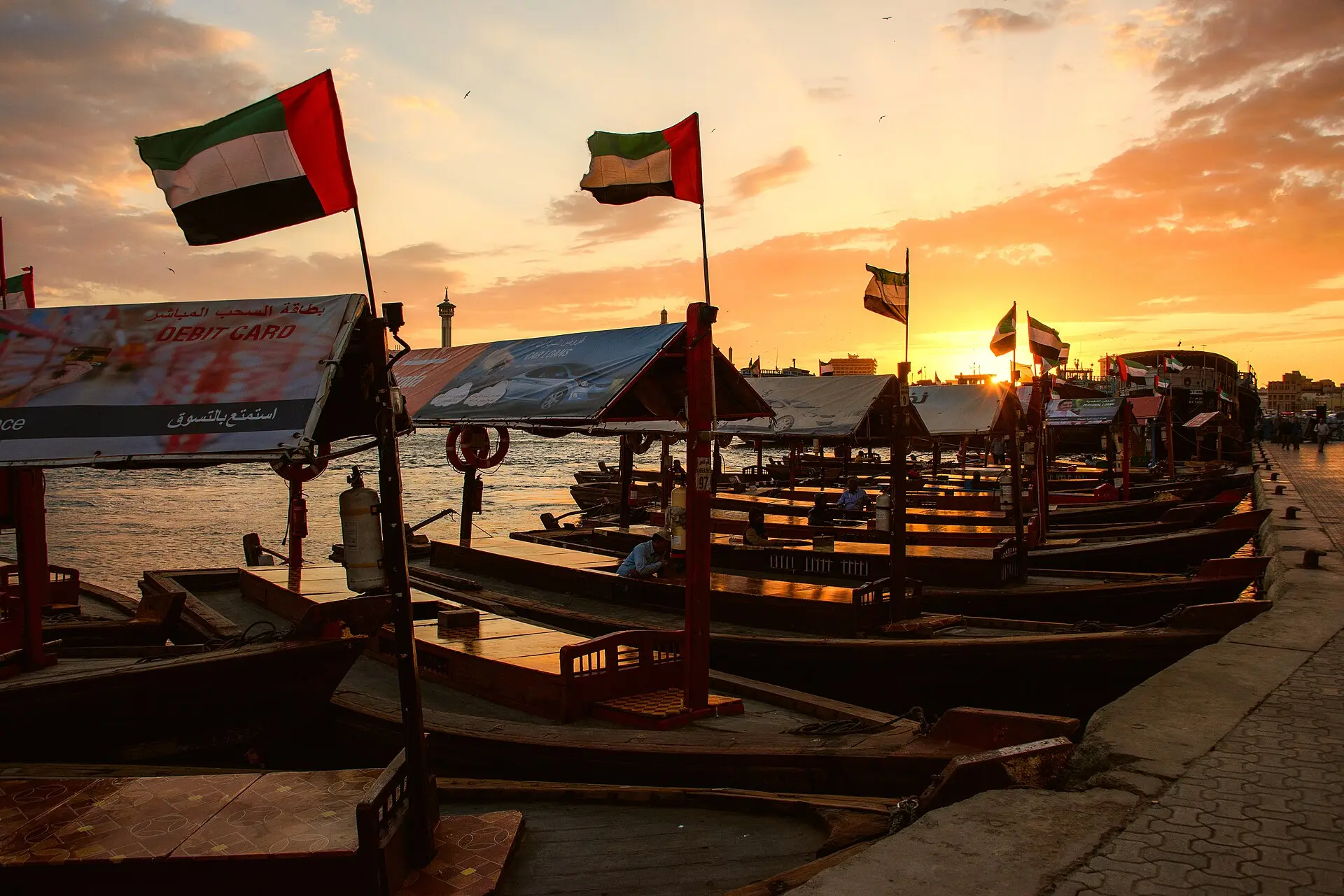 Vertäute hölzerne Abra-Boote mit Stoffdächern und VAE-Flaggen am Dubai Creek, rechts die Uferpromenade, dahinter orangefarbener Abendhimmel
