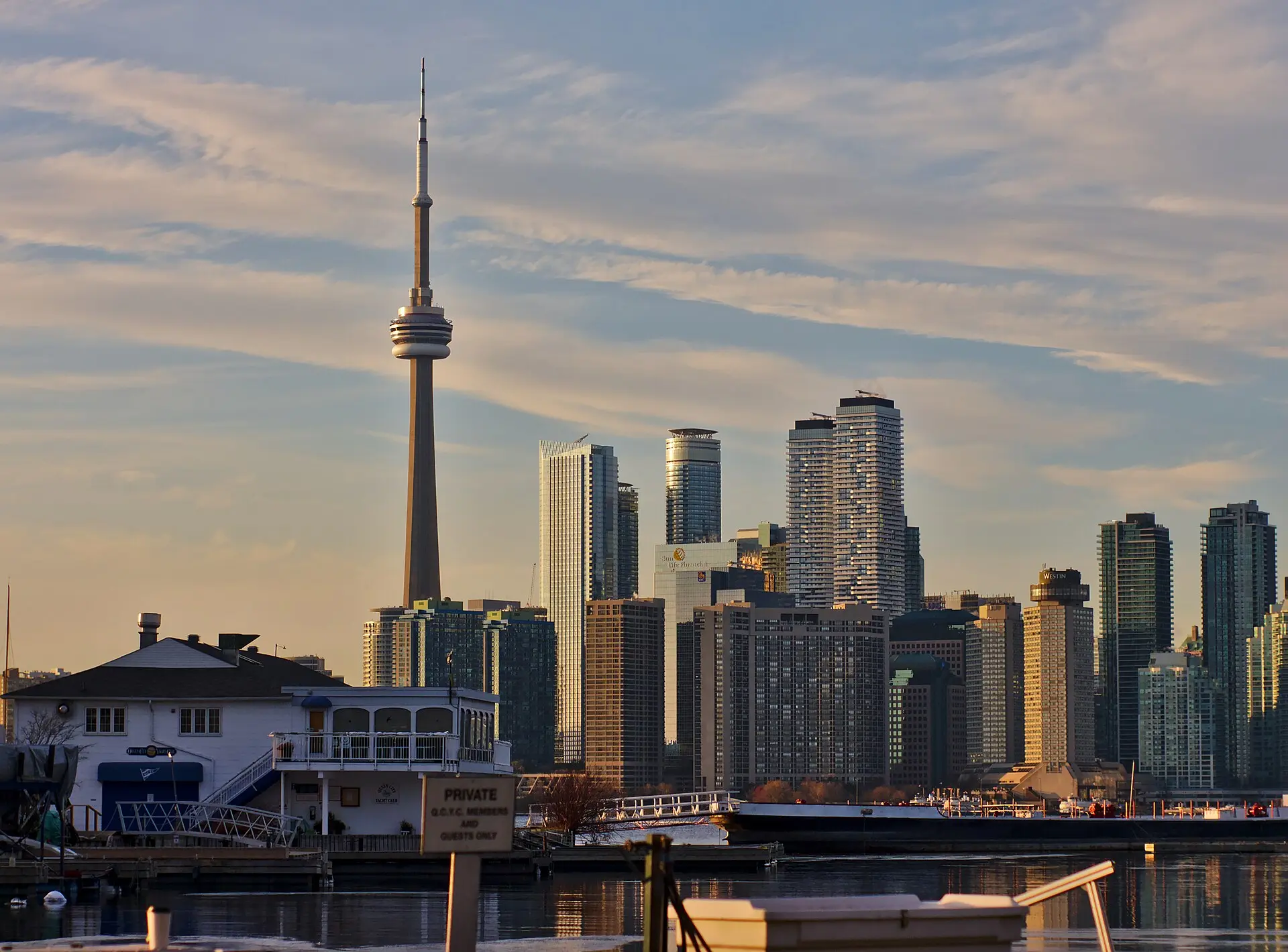Skyline von Toronto mit dem CN Tower im Licht der tief stehenden Sonne, davor ein weißes Clubhaus auf den Toronto Islands und ruhiges Wasser