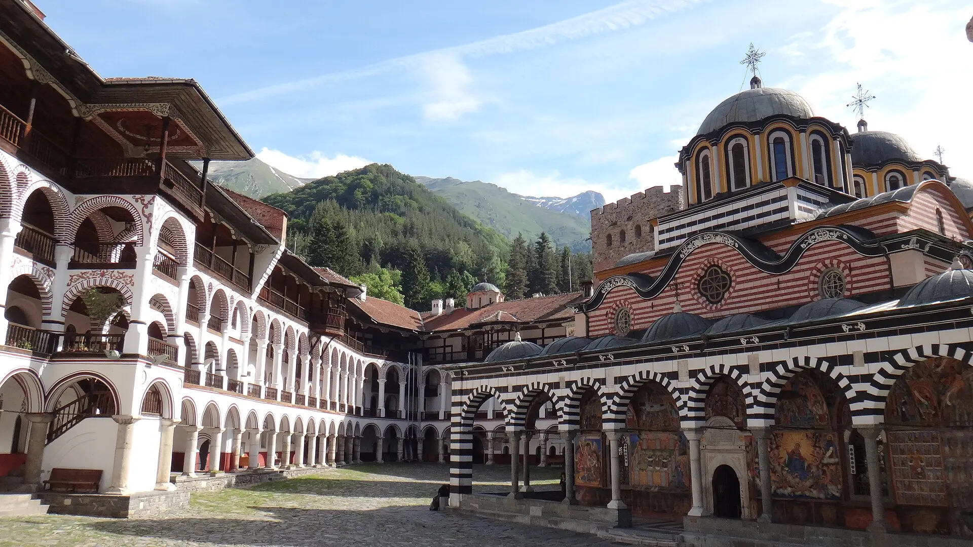 Innenhof des Rila-Klosters: links Wohntrakt mit weißen Bogengängen und Holzbalkonen, rechts die Hauptkirche, dahinter bewaldete Berge