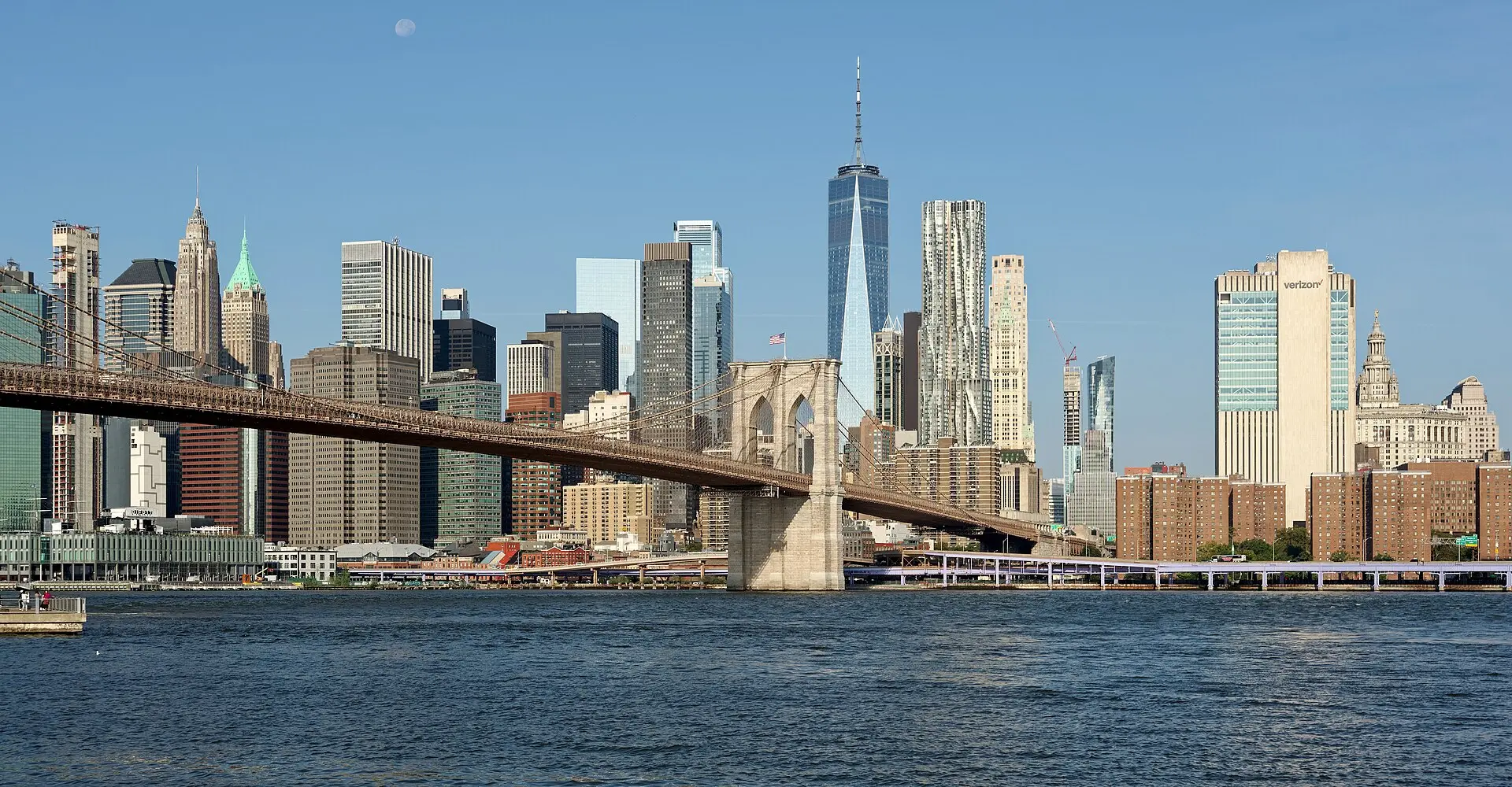 Brooklyn Bridge über dem East River, dahinter die Hochhäuser von Lower Manhattan mit One World Trade Center unter blauem Himmel