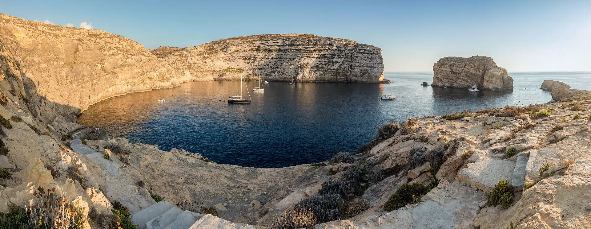 Dwejra-Bucht auf Gozo im Abendlicht mit Booten, hellen Kalksteinklippen und einem freistehenden Felsblock im Meer