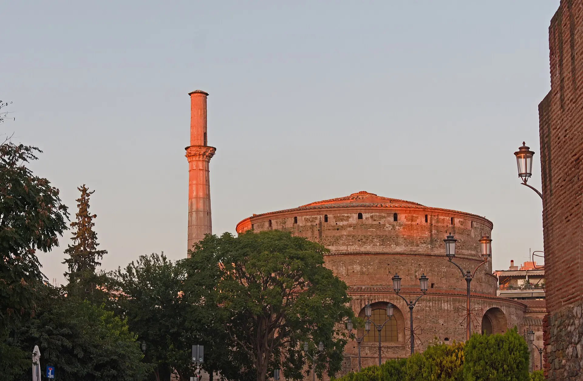 Rundbau der Rotunde mit Minarett im Abendlicht, davor Bäume und Laternen, rechts die Ziegelmauer des Galeriusbogens