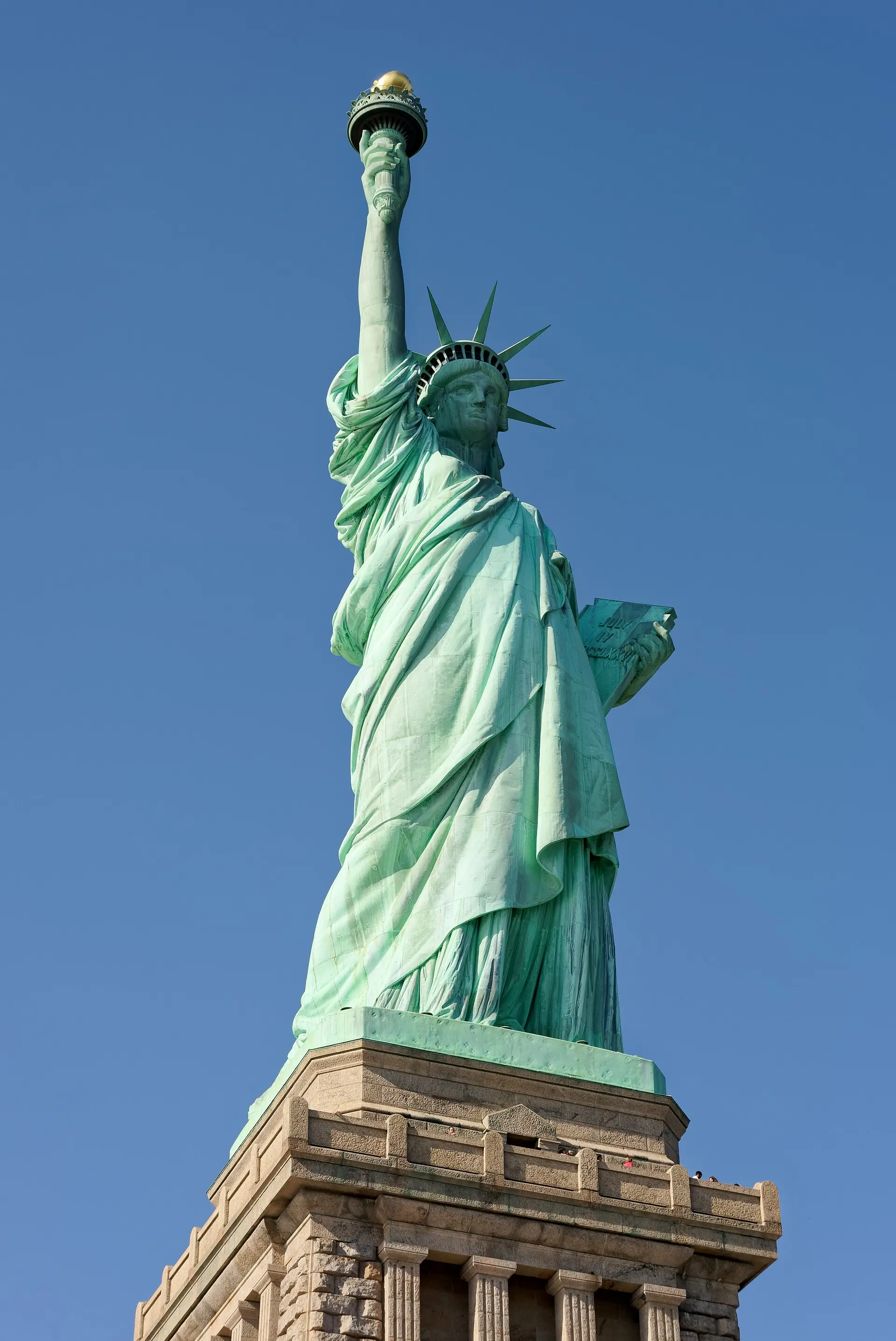 Freiheitsstatue von schräg unten vor wolkenlosem blauem Himmel, mit Fackel, Krone und Tafel, darunter der Granitsockel
