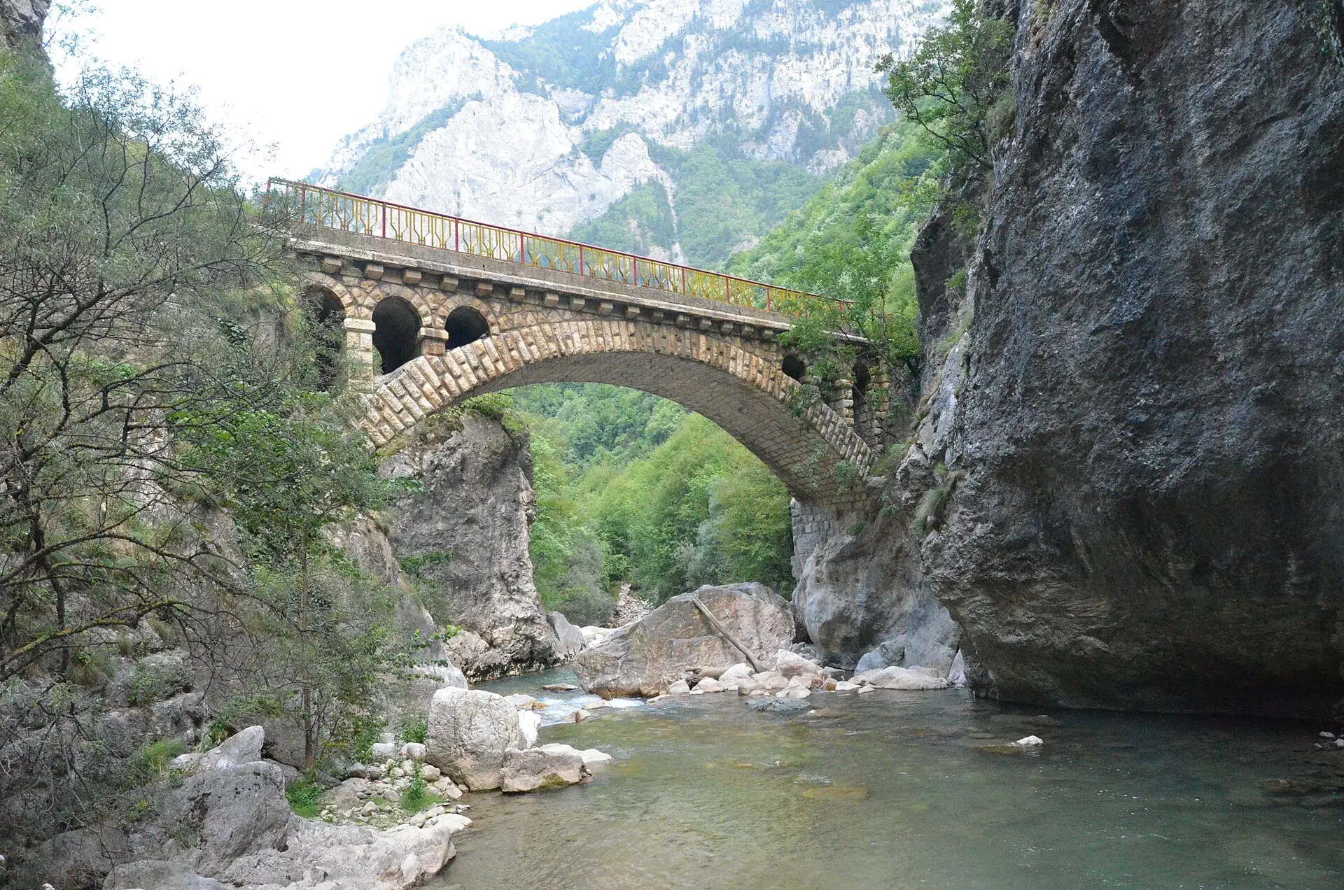 Steinerne Bogenbrücke mit rotem Geländer über dem Fluss in der Rugova-Schlucht, rechts eine steile Felswand, im Flussbett helle Felsblöcke