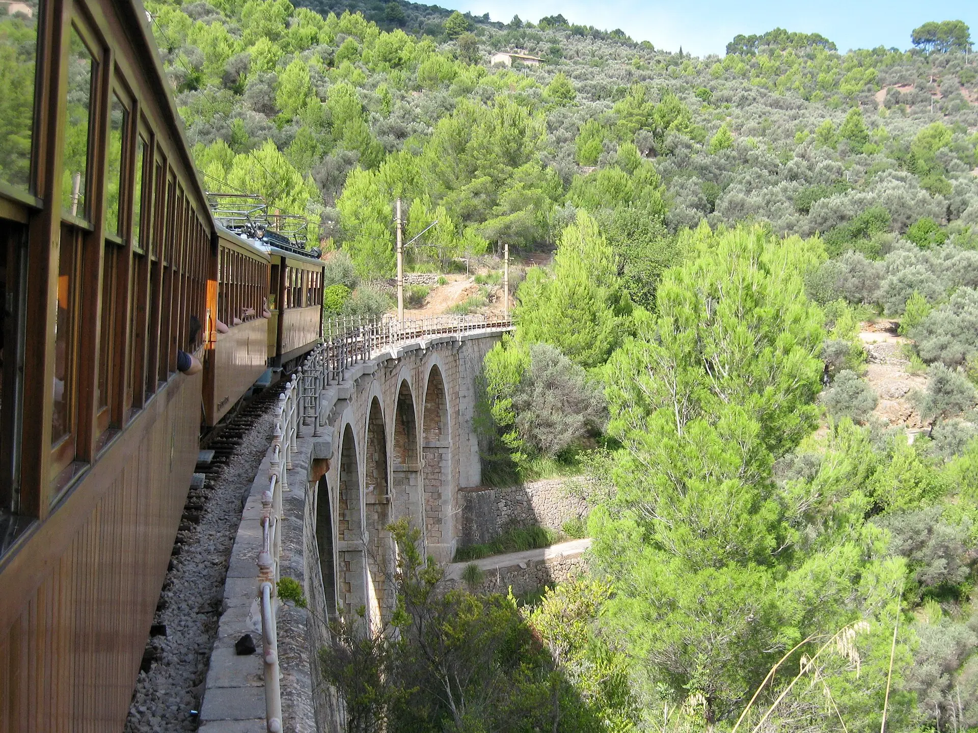 Holzwaggons des Tren de Sóller in einer Kurve auf einem gemauerten Bogenviadukt, rechts Hänge mit Pinien und Olivenbäumen