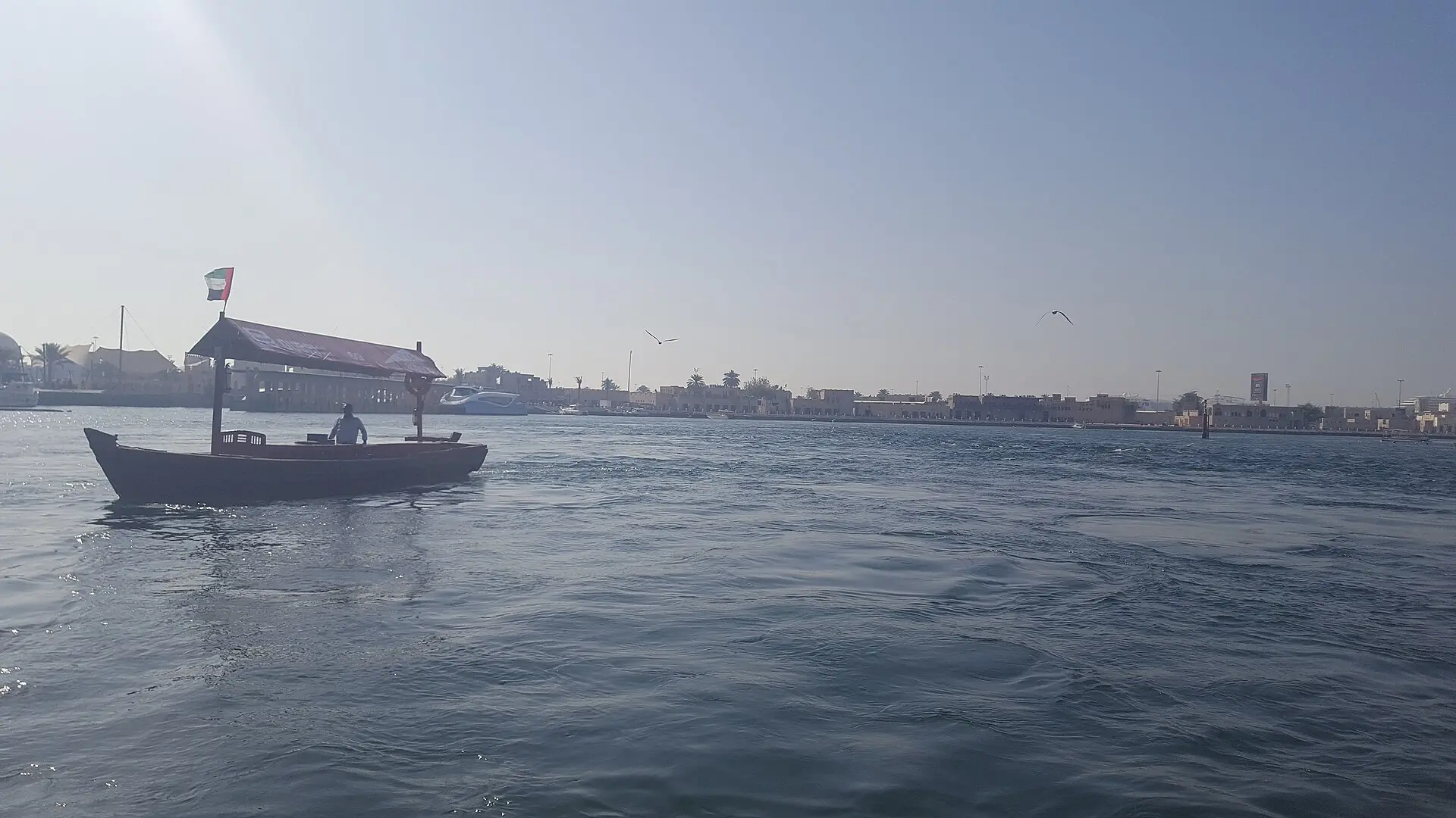 Holzabra mit rotem Sonnendach und VAE-Flagge auf dem Dubai Creek, am Ufer flache sandfarbene Häuser und Palmen, darüber zwei Vögel