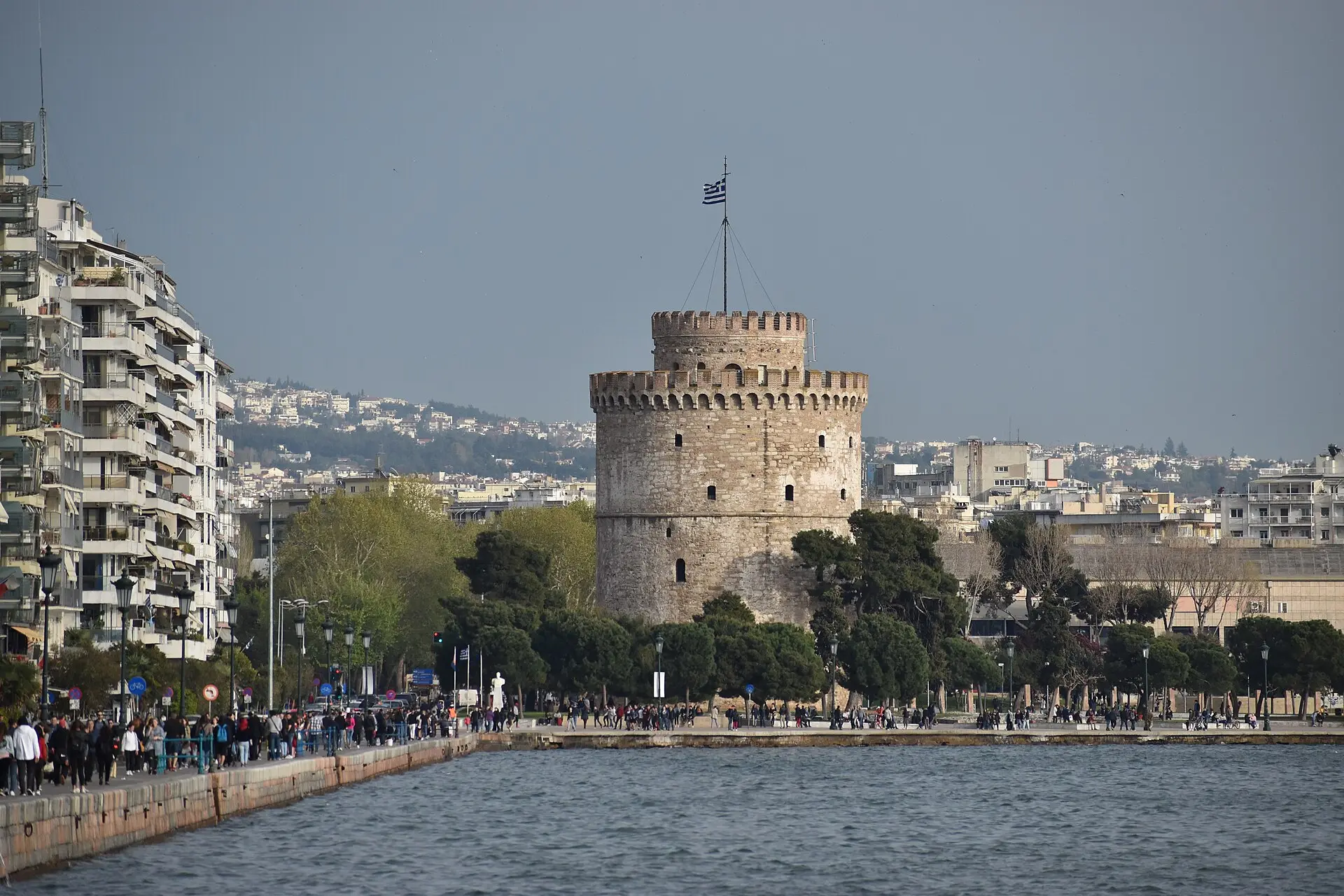 Weißer Turm von Thessaloniki mit griechischer Flagge, über das Wasser gesehen, links Wohnhäuser und viele Spaziergänger am Ufer