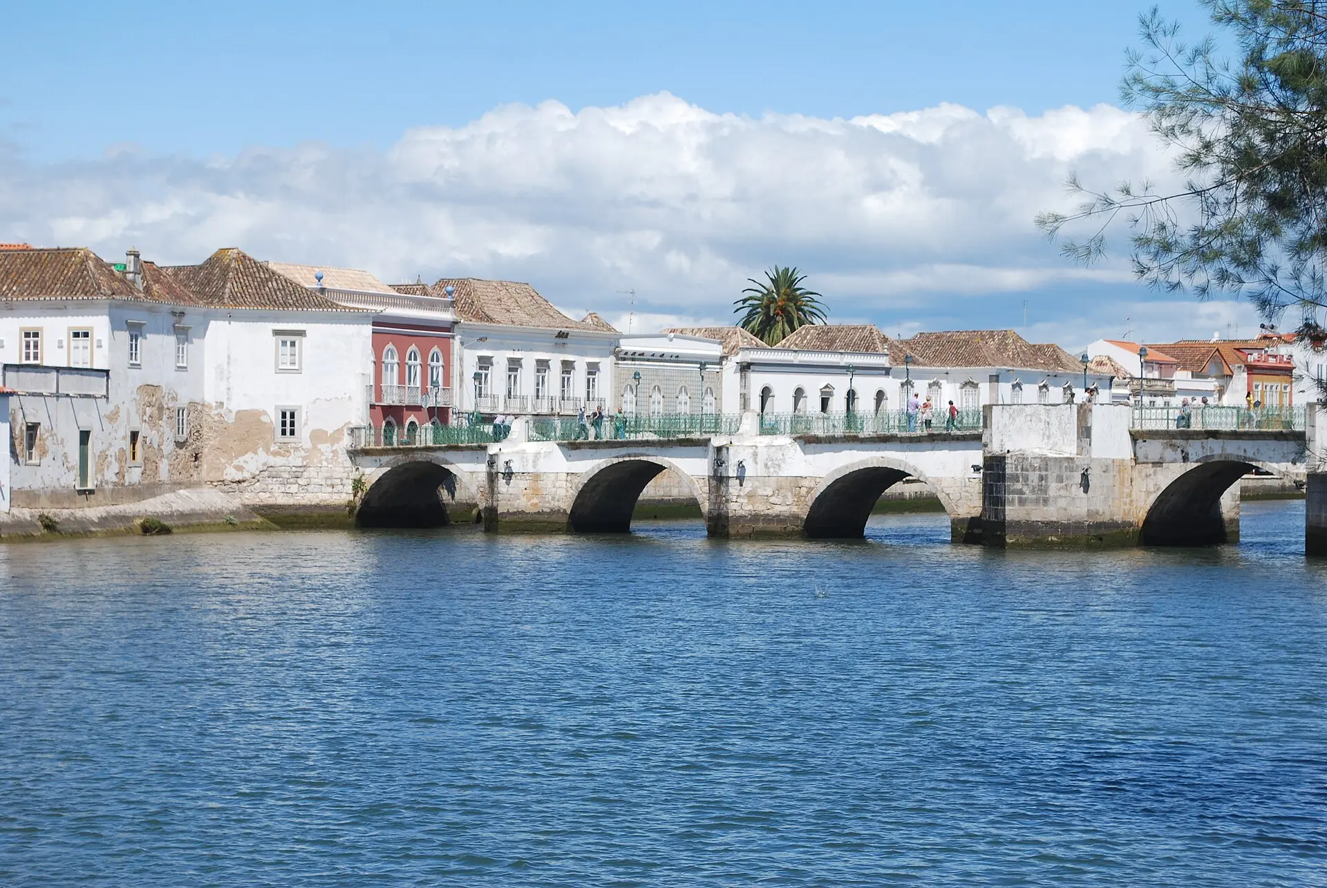 Die Steinbögen der Ponte Antiga über den blauen Rio Gilão in Tavira, dahinter weiße Häuser mit Ziegeldächern und eine Palme