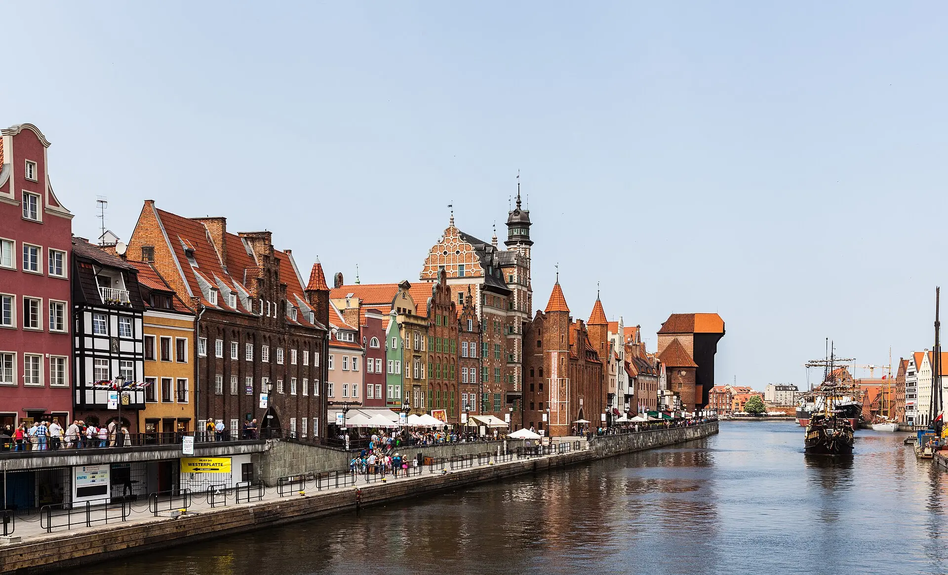 Bunte Giebelhäuser am Motława-Kai in Danzig mit dem dunklen Krantor, auf dem Fluss ein Segelschiff im alten Stil