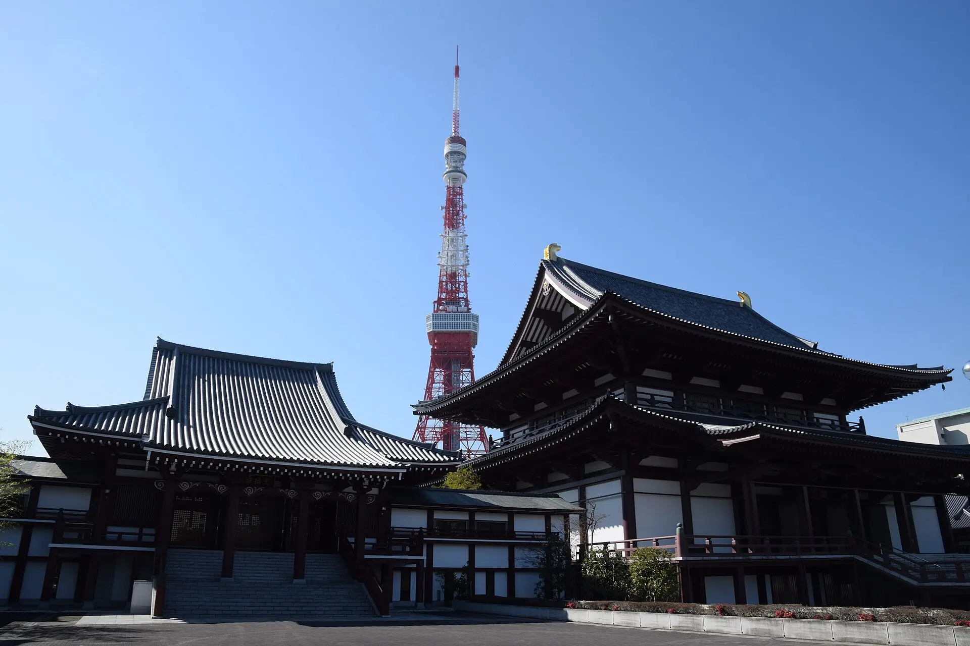 Zwei Hallen des Tempels Zojo-ji mit geschwungenen Dächern, dazwischen ragt der rot-weiße Tokyo Tower in den blauen Himmel